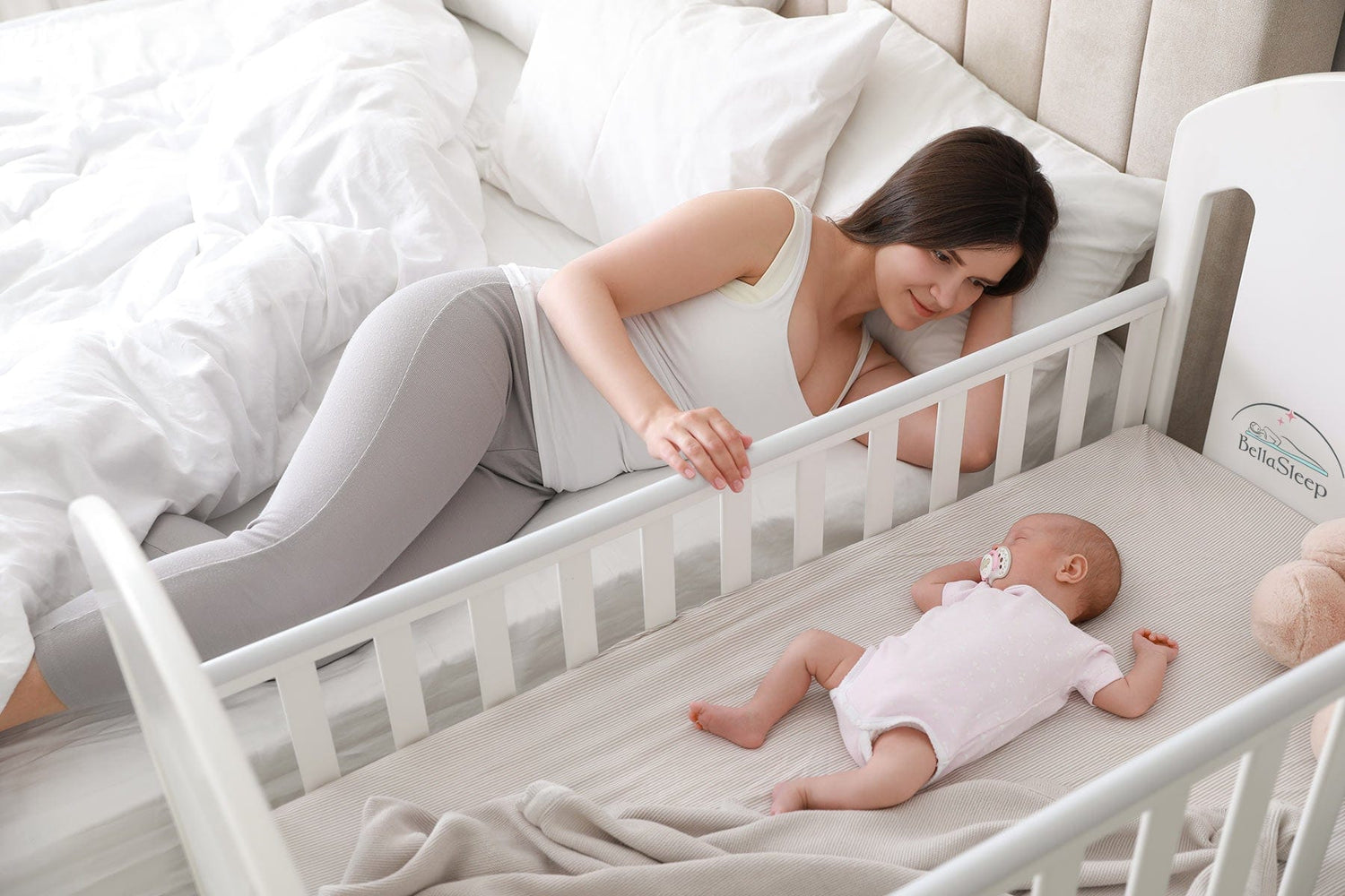 Mutter liegt neben dem Babybett auf ihrem Bett und berührt liebevoll ihr Baby – Nähe und Geborgenheit mit BellaSleep Baby Matratze