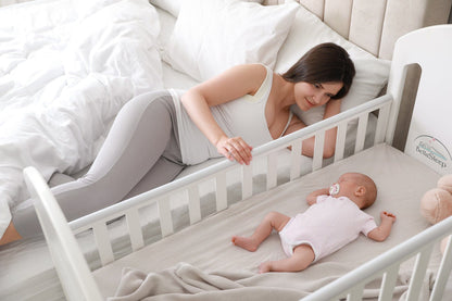Mutter liegt neben dem Babybett auf ihrem Bett und berührt liebevoll ihr Baby – Nähe und Geborgenheit mit BellaSleep Baby Matratze